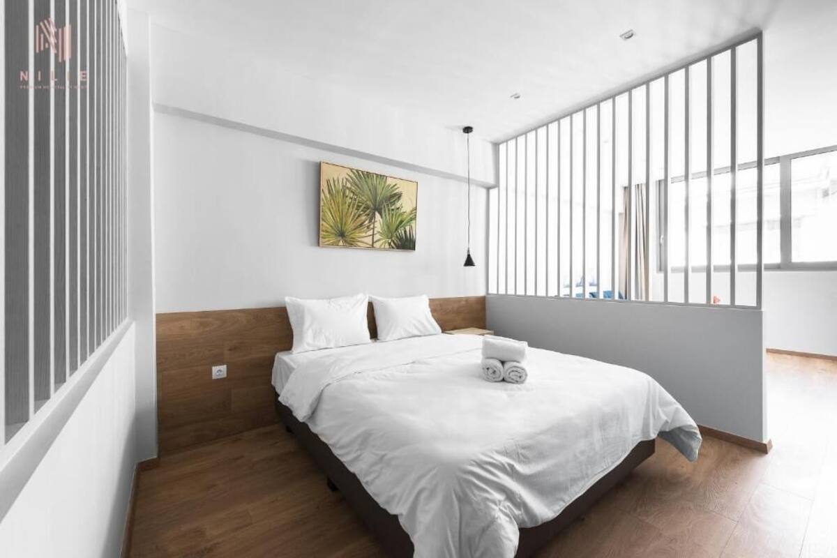 A modern bedroom with a large bed, a wooden headboard, a wall-mounted artwork, and a large window with vertical blinds.