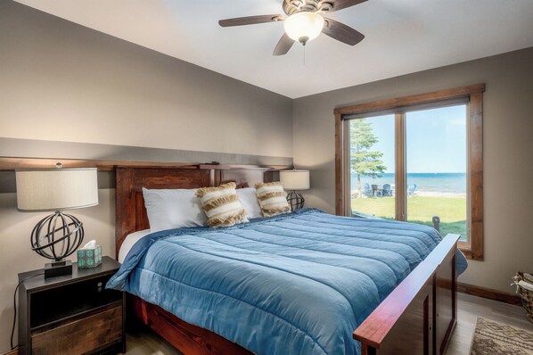 A bedroom with a large bed, a wooden headboard, a nightstand with a lamp, and a window with a view of the beach. - Luxury lakefront home - 6 BRs, kids, pets, EVs, elevator - family celebrations (Baileys Harbor)