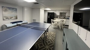 A room with a ping pong table, a sofa, a dining table with chairs, and a kitchen area with cabinets and appliances.