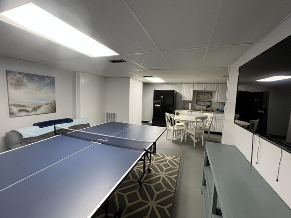 A room with a ping pong table, a sofa, a dining table with chairs, and a kitchen area with cabinets and appliances. - Lake Ontario Beach House Getaway-House #2 (Henderson)