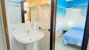 Un baño con lavamanos, espejo y ducha. En la habitación contigua hay una cama con manta azul.