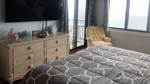 A bedroom with a large bed, a television mounted on the wall, a wicker dresser, and a chair by the window. - The Plaza - Direct Oceanfront  Luxury Condo with Beach bar-Discount weekly  (Ocean City)
