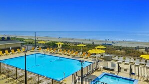 A swimming pool with sun loungers and umbrellas, a beach in the background, and a clear blue sky. - The Plaza - Direct Oceanfront  Luxury Condo with Beach bar-Discount weekly  (Ocean City)