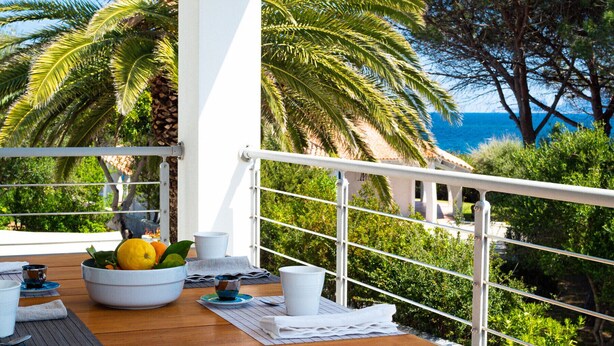 A balcony with a wooden table, white chairs, a bowl of fruit, and a view of the sea and palm trees.