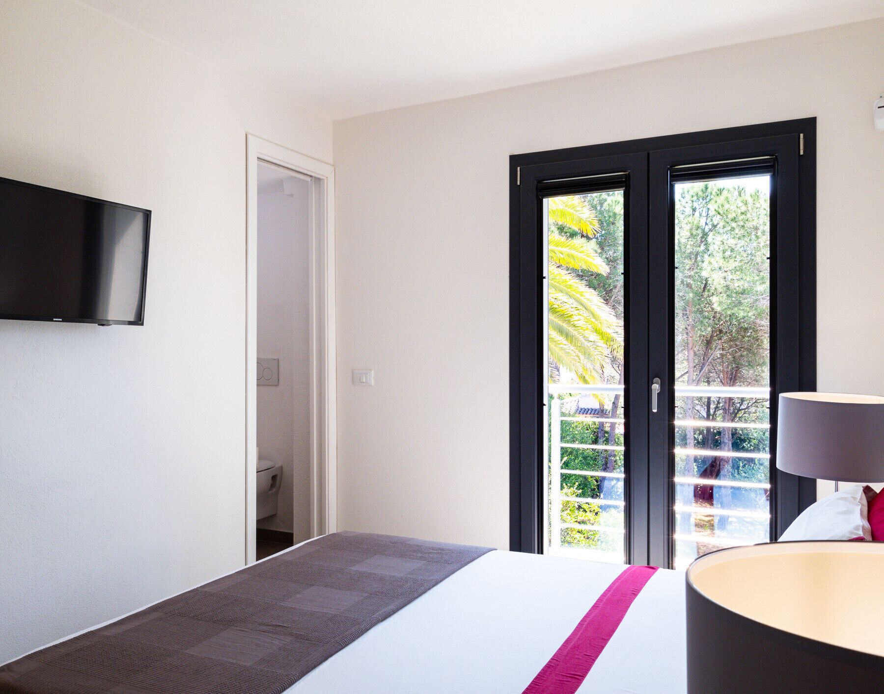 Une chambre d’hôtel avec un lit, un téléviseur fixé au mur, une porte donnant sur un balcon avec vue sur des arbres et un palmier, et une lampe sur la table de chevet.