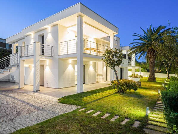 A modern two-story house with a spacious front yard, a staircase leading to the entrance, and a well-lit balcony.