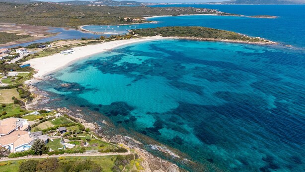 Luftaufnahme einer Küstenregion mit Sandstrand, klarem blauen Wasser und verstreuten Gebäuden.