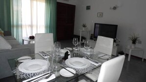 A dining area with a glass table set for four, surrounded by white chairs. There is a sofa and a small round table with a plant and a fruit bowl in the background.