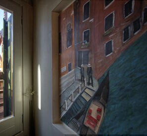 A view from a window of a rooftop terrace with a railing, a vase with flowers, and a painting of a street scene.