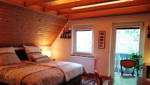 Een gezellig slaapkamer met houten plafond, een groot bed met beddengoed met patroon, een nachtkastje met lamp, een raam met uitzicht op groen, en een eethoek met tafel en stoelen.