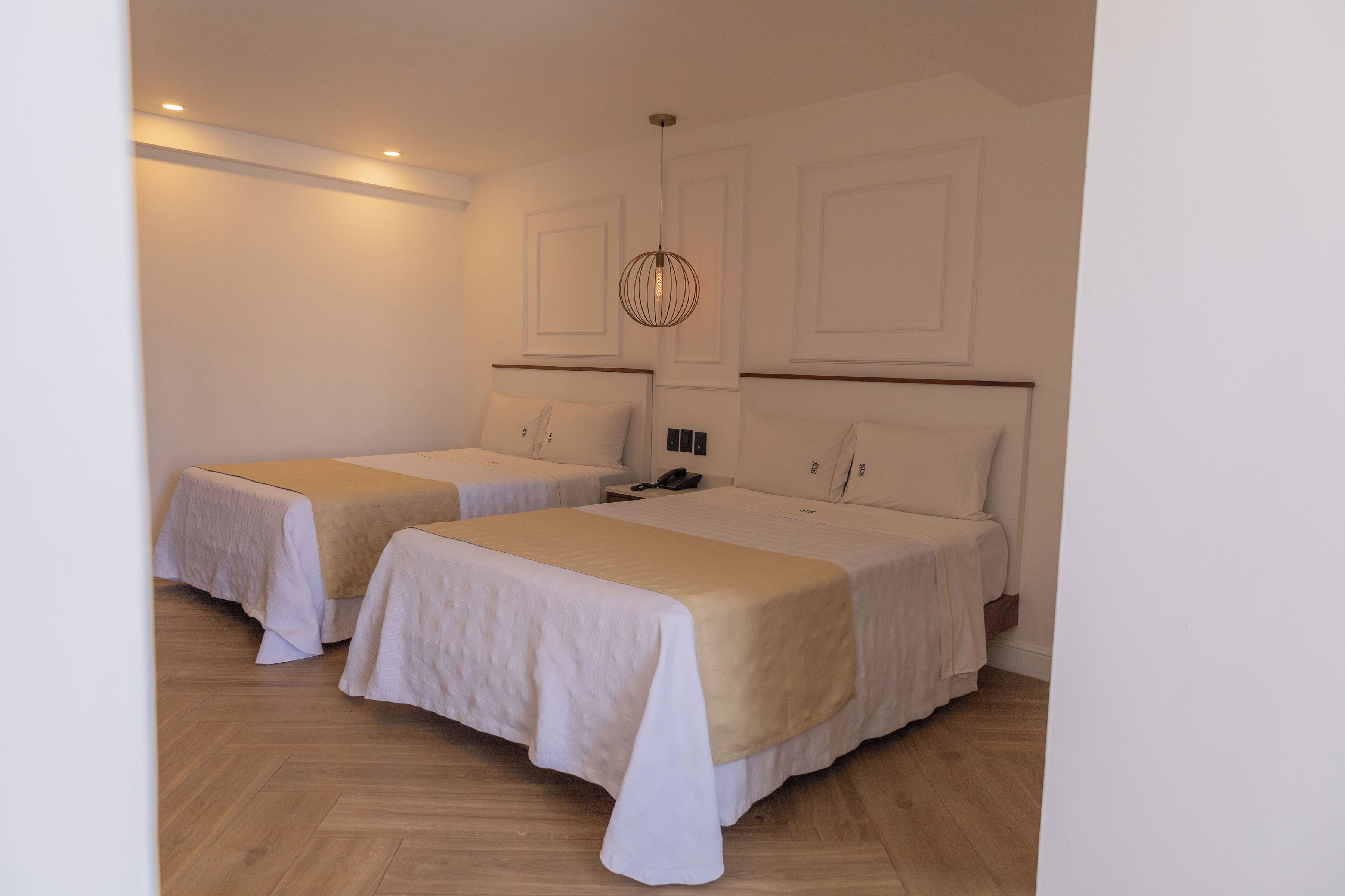 A hotel room with two beds, each with a white bedspread and a beige throw. There is a wooden floor and a hanging light fixture above the beds.