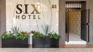 Letrero de SIX HOTEL con diseño de pared de mármol, el número 85 y dos macetas negras con diversas plantas al frente.