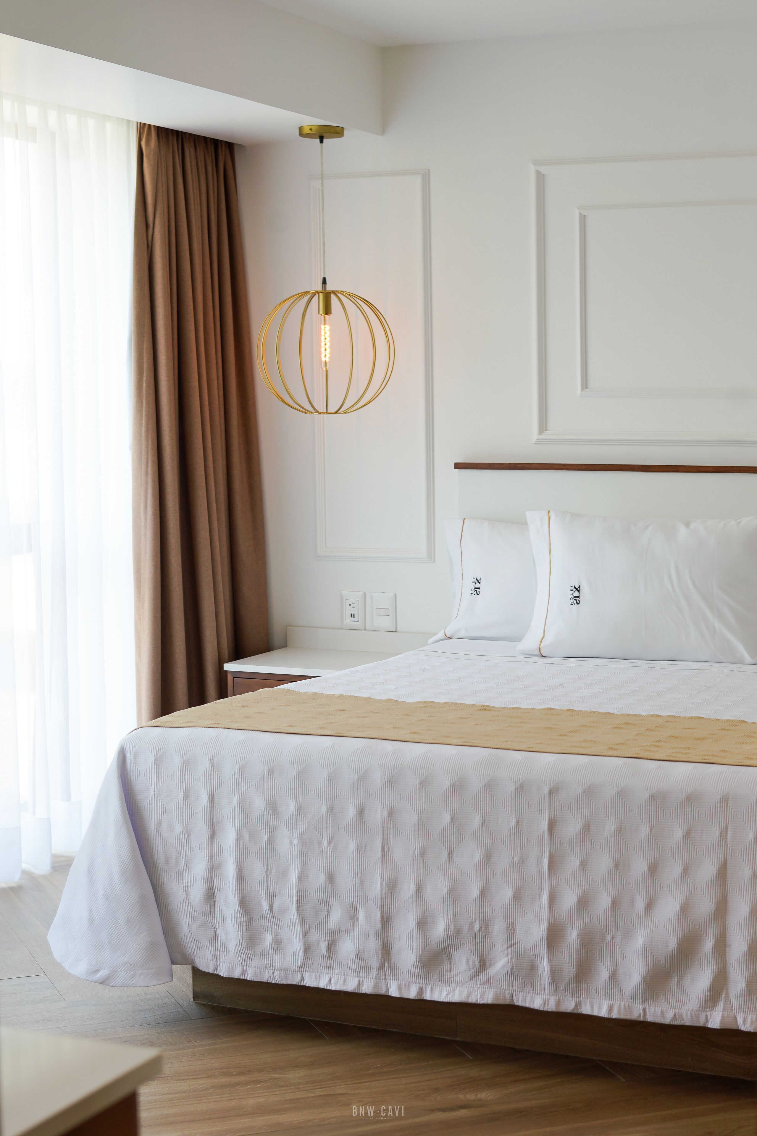Una cama bien hecha con sábanas blancas, cabecera de madera y lámpara colgante.