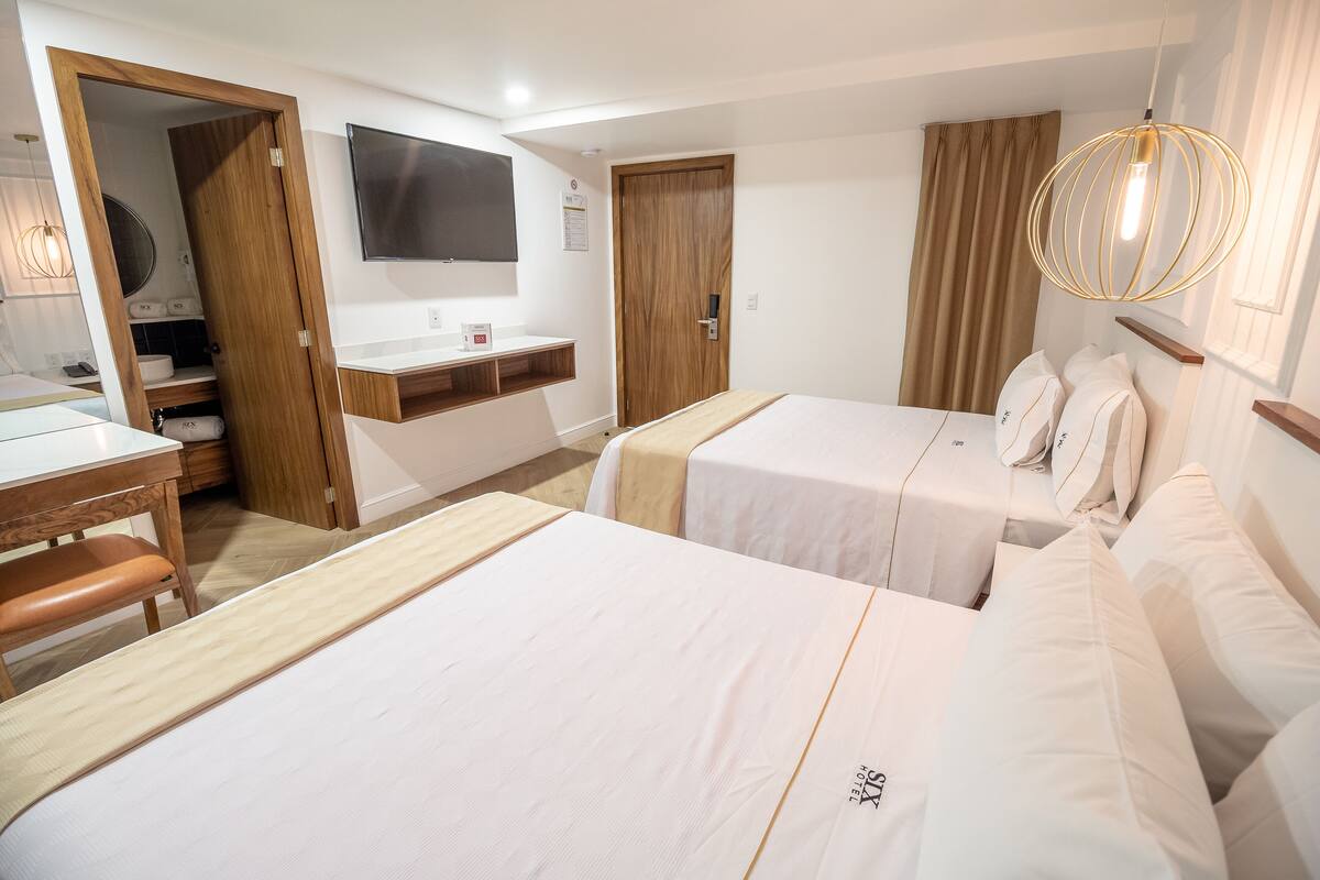 Habitación de hotel con dos camas, un escritorio de madera, un baño pequeño y un televisor montado en la pared.