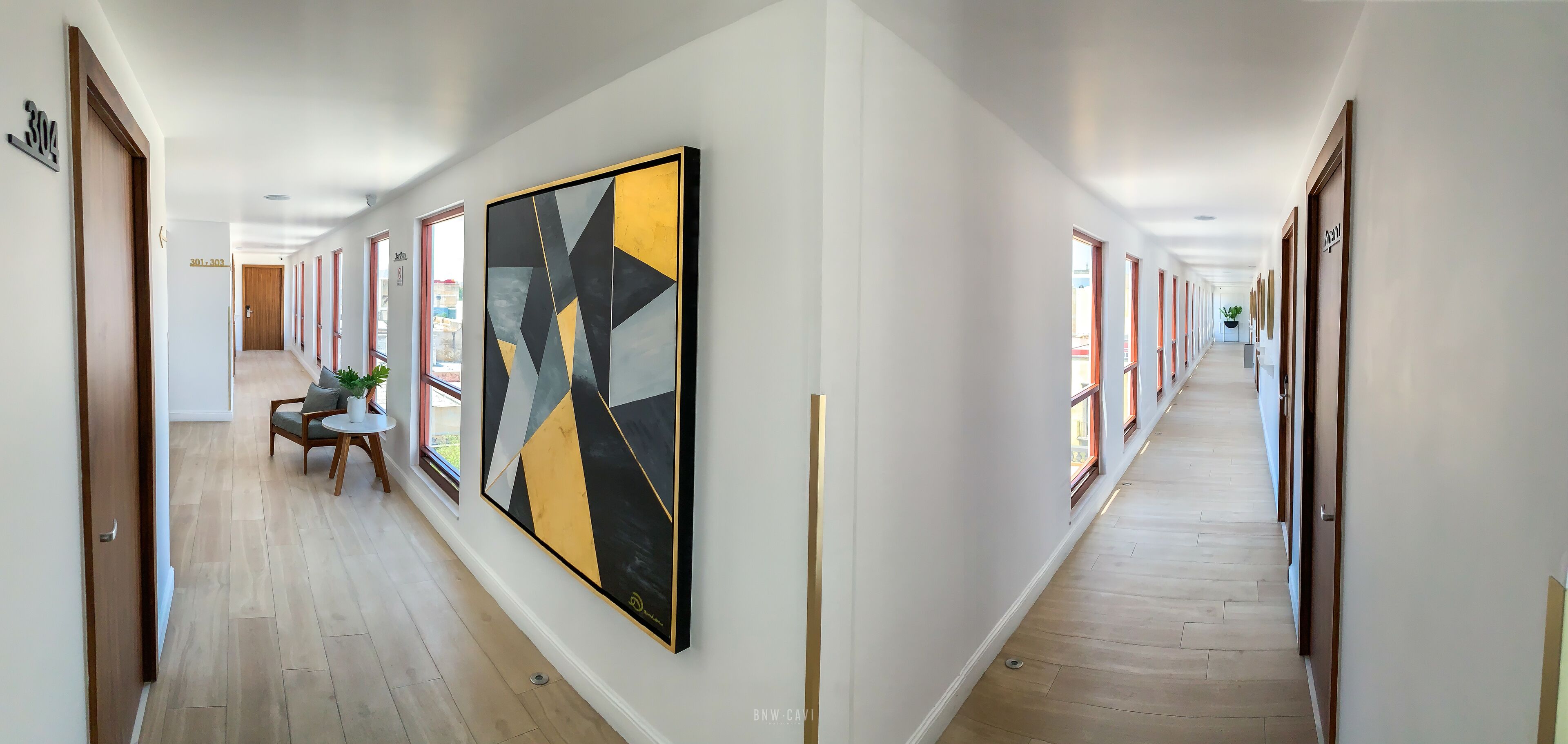 A long, narrow hallway with wooden floors and white walls, featuring a large abstract painting on one wall and a small seating area with a table and chairs.