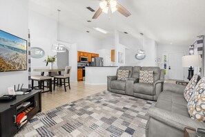 A modern living room with a gray sofa, a dining area with a wooden table, and a kitchen with wooden cabinets.