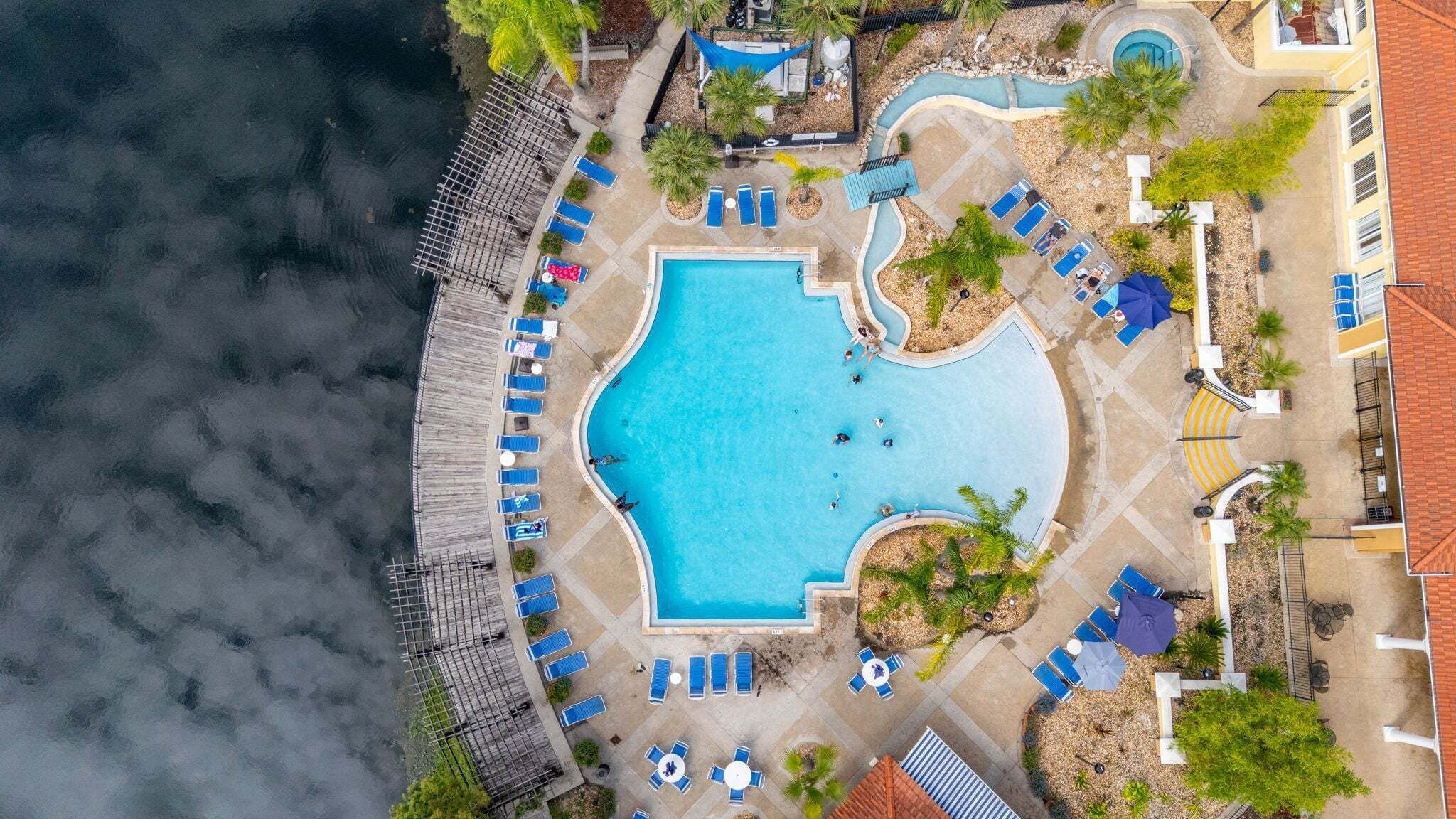 An aerial view of a resort with a central swimming pool, sun loungers, and a wooden deck overlooking the water.