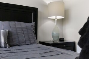 A bed with grey bedding and a black headboard, a bedside table with a lamp, and a small drawer.