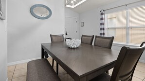 Ein Esszimmer mit einem rechteckigen Tisch, vier Stühlen, einer weißen Dekorationsvase, einem runden Spiegel und einer Deckenleuchte.