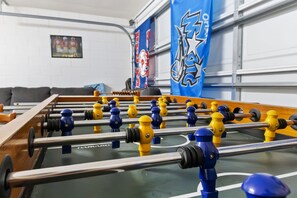 Ein Fußballtisch mit Holzrahmen und Metallstangen, platziert in einem Raum mit weiß verputzter Backsteinwand und einer blauen Flagge mit Sternenmuster.