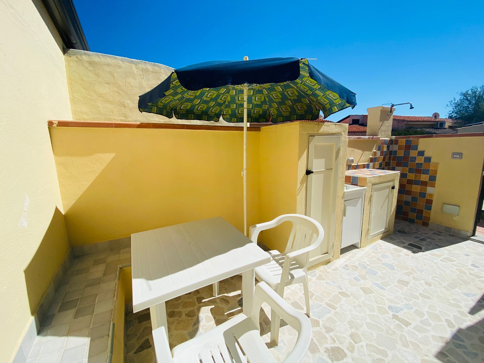 A small outdoor patio with a white table and chairs, a green and black umbrella, and a tiled wall.