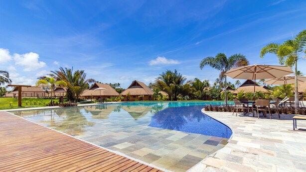 A swimming pool with a wooden deck and stone tiles, surrounded by palm trees and umbrellas. - Apto Completo no Eco Resort (Rio Formoso)