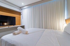 Habitación de hotel con una cama grande, un televisor de pantalla plana montado en un panel de madera, cortinas blancas y una lámpara de noche.
