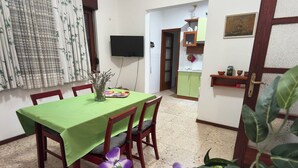 A dining area with a green table, wooden chairs, a flat-screen TV, a kitchen in the background, and a door with a decorative handle.