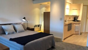 A bedroom with a bed, bedside table, lamp, and a view of a kitchenette.