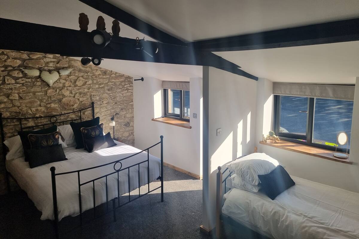 Un dormitorio con pared de piedra, cama con cabecero oscuro, ventana con vistas y una pequeña mesa con lámpara.
