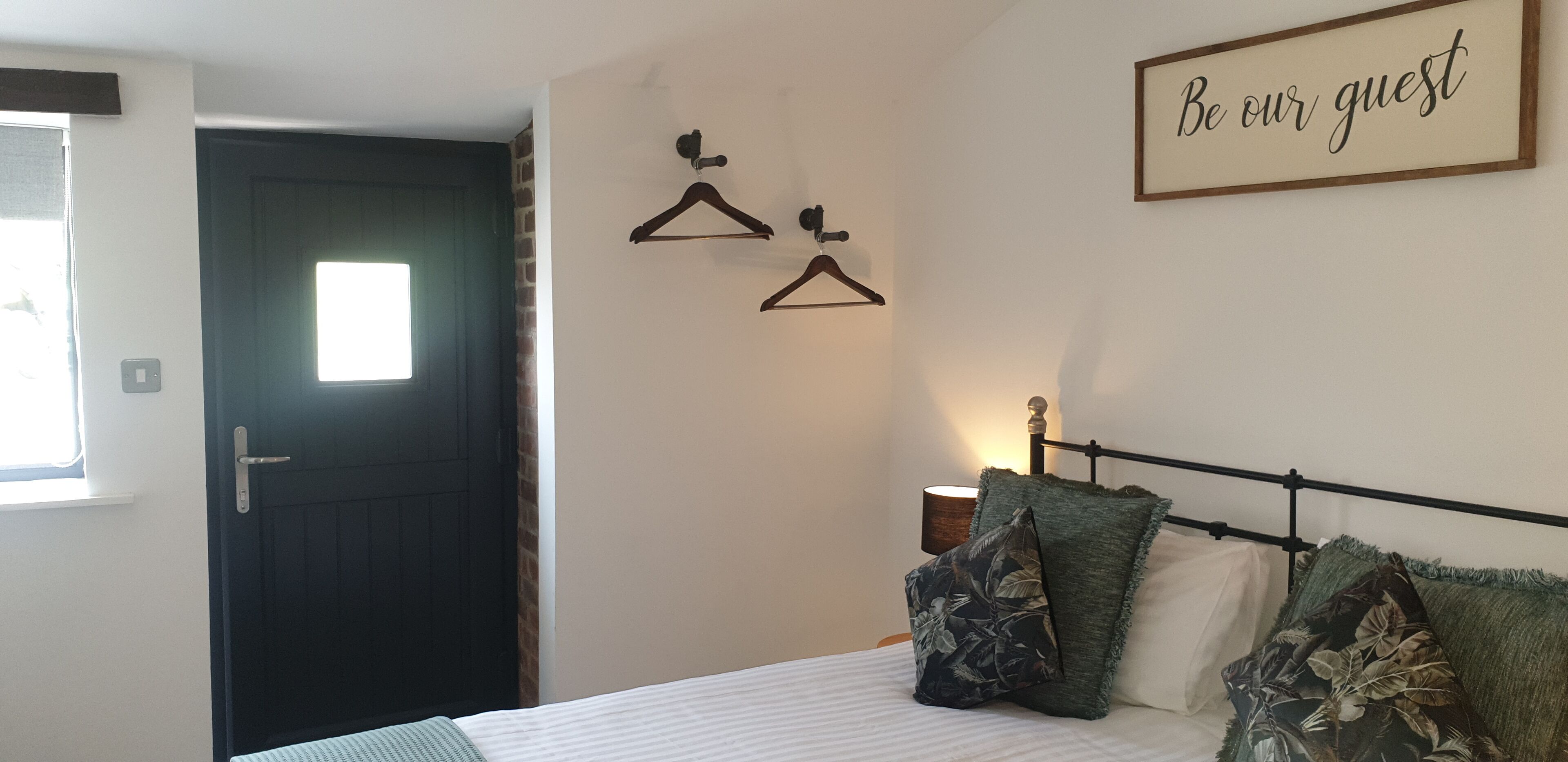 A bedroom with a bed, a wall-mounted light, a door, and a sign that says "Be our guest".
