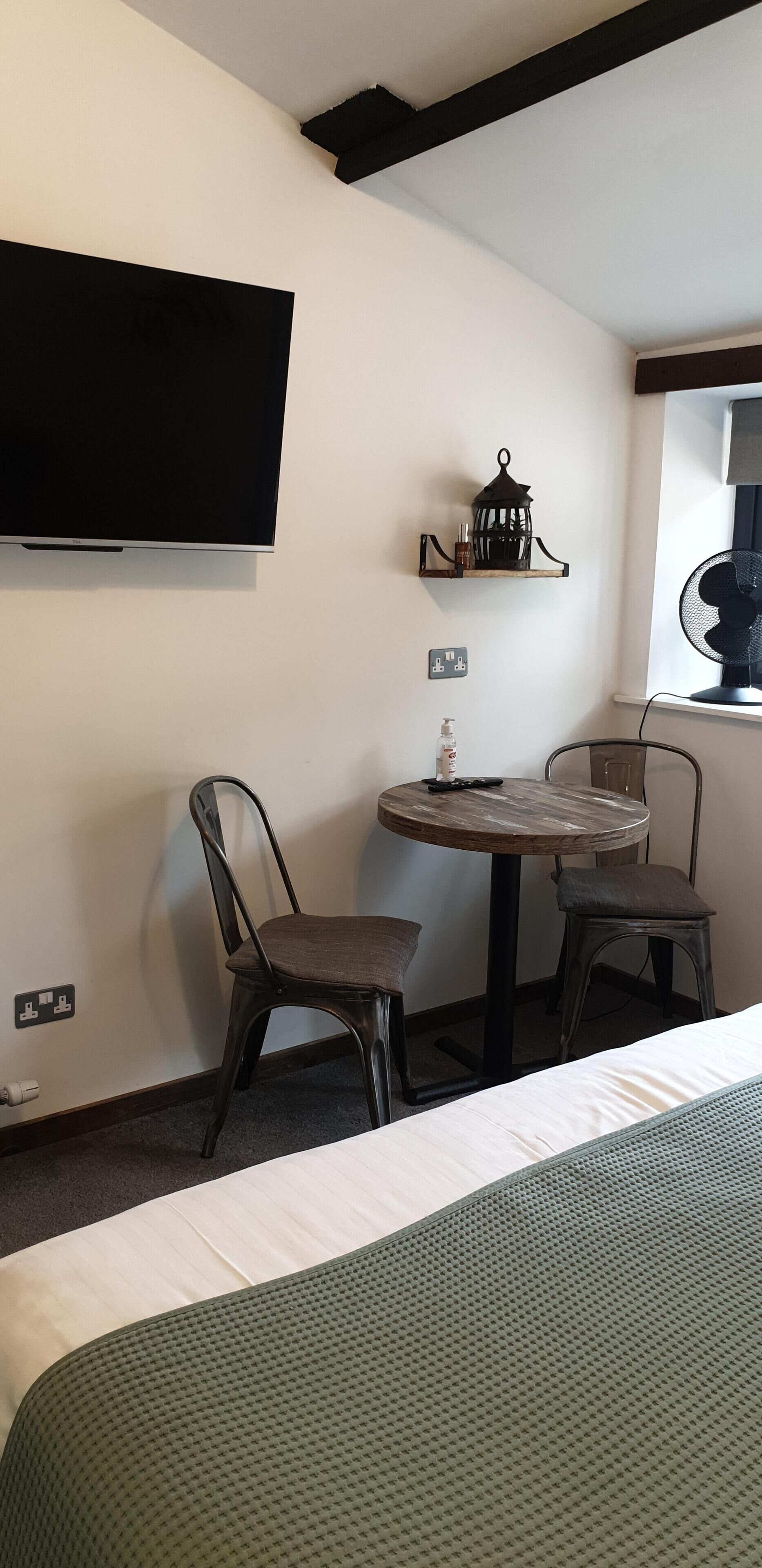 Een kamer met een bed, een tafel met twee stoelen, een aan de muur bevestigde televisie, een klein plankje met een lantaarn, en een raam met een ventilator.