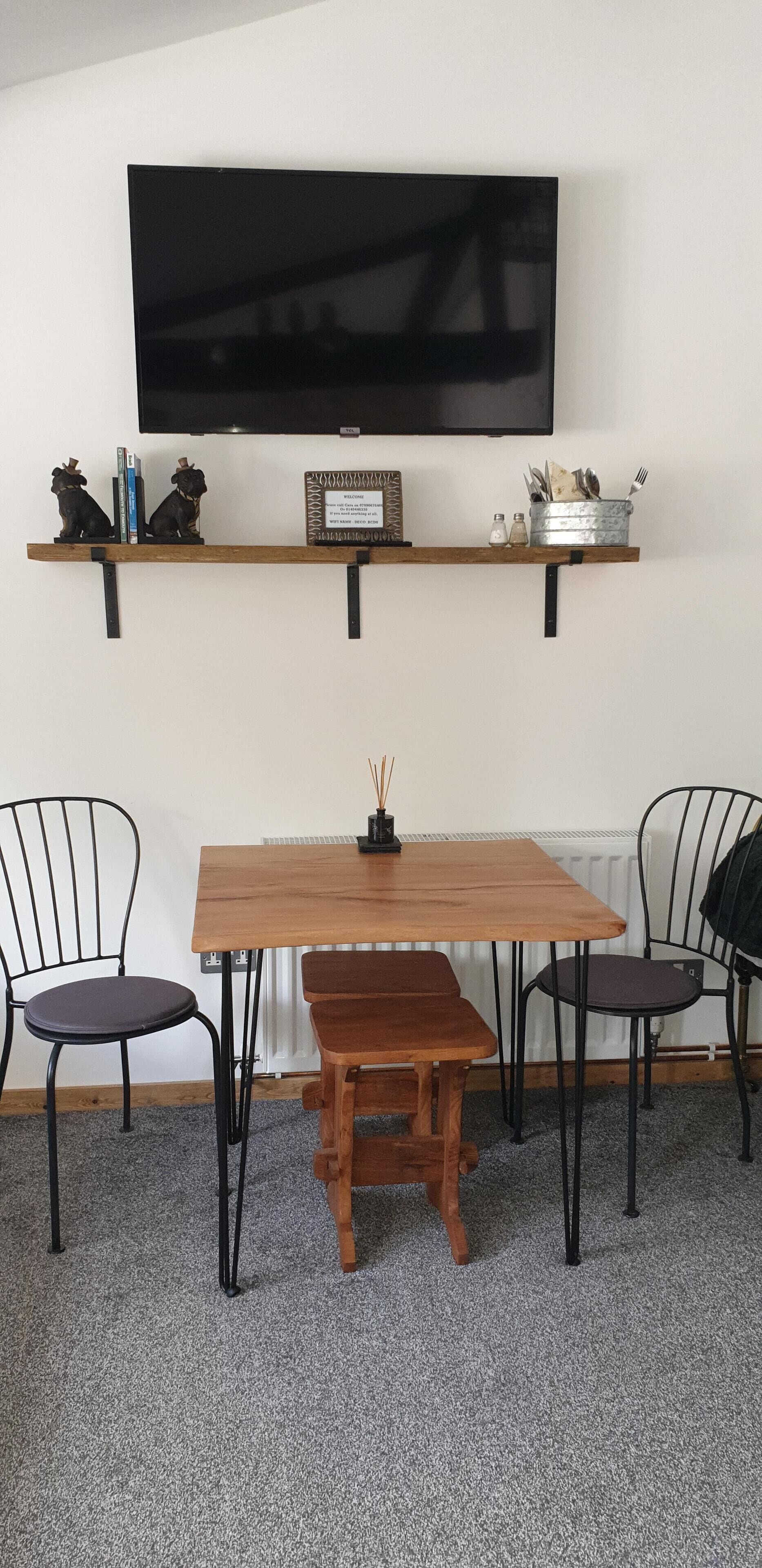 Un comedor con una mesa de madera, dos sillas, un banco, un televisor montado en la pared y un estante con objetos decorativos y utensilios de cocina.