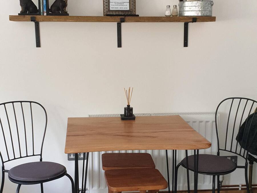 Un comedor con una mesa de madera, dos sillas, un banco, un televisor montado en la pared y un estante con objetos decorativos y utensilios de cocina.