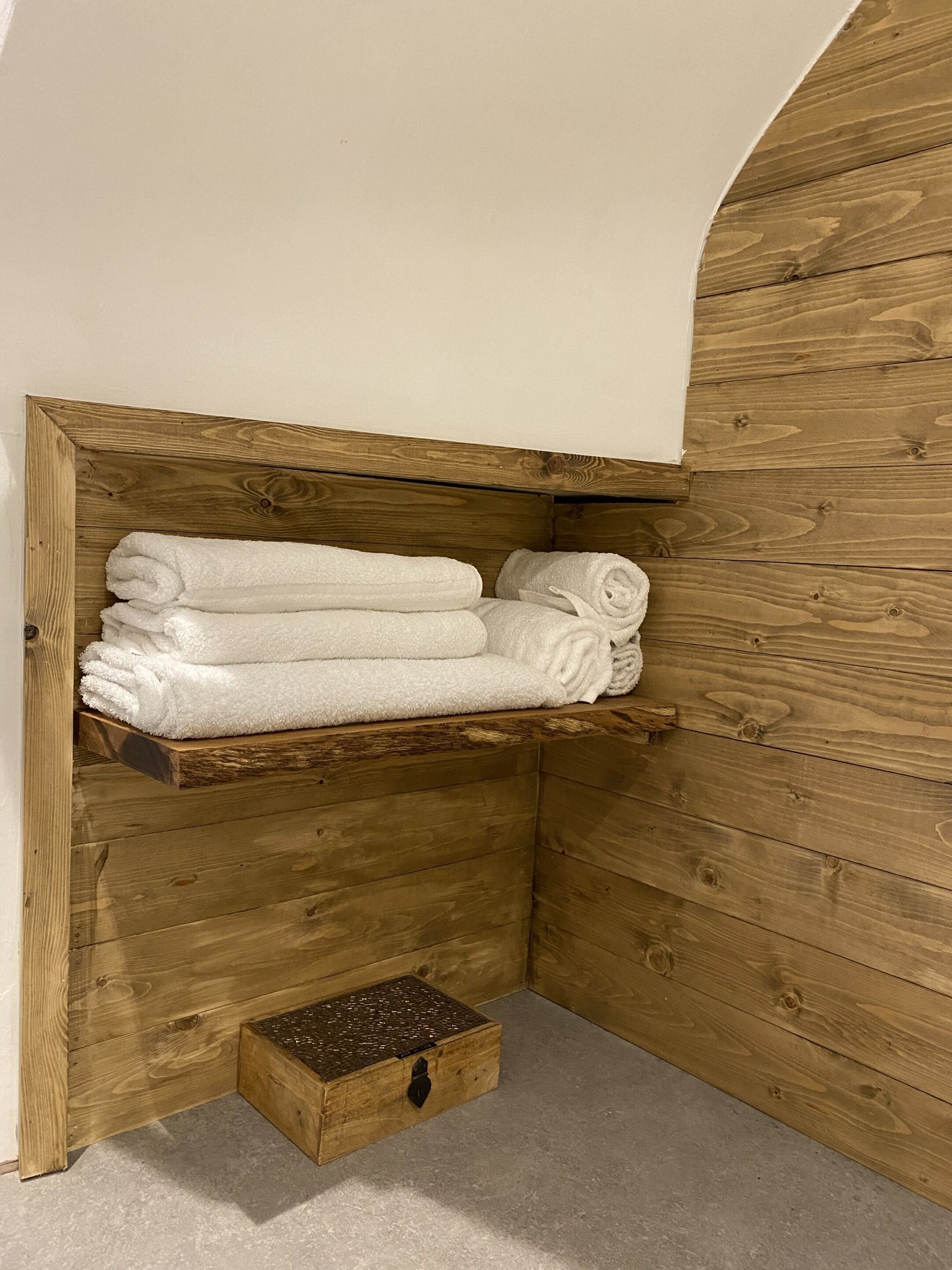 A corner with a wooden shelf holding folded towels and a small chest of drawers.