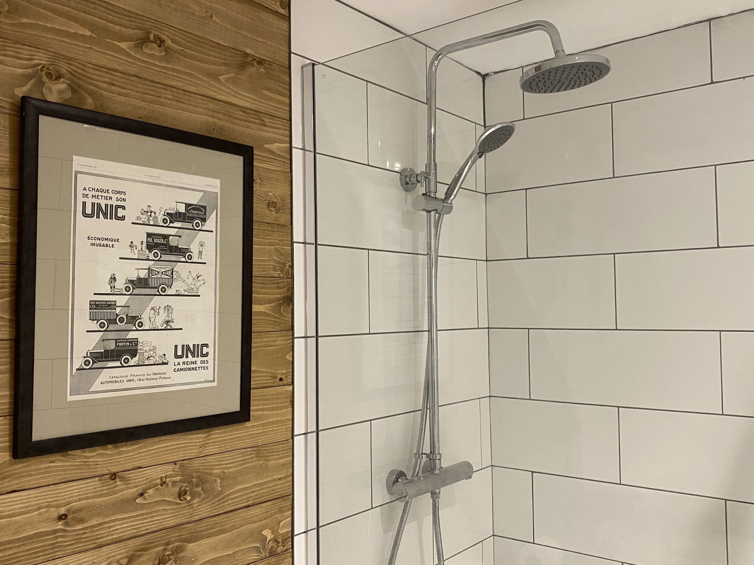 En inramad reklam för UNIC på en vägg av trä, och en dusch med ett duschmunstycke som kan hållas i handen och en väggmonterad blandare.
