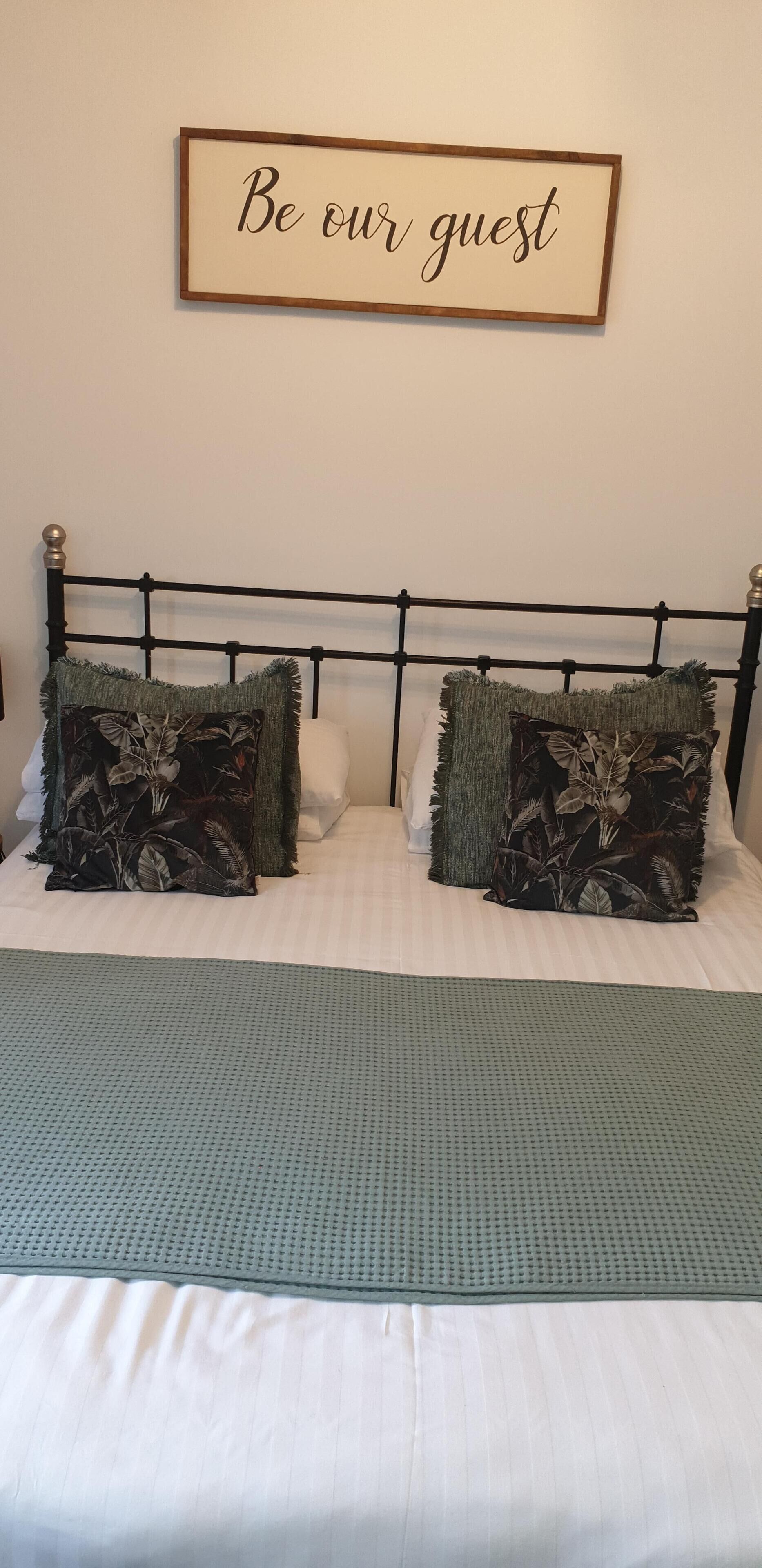 Un lit bien fait, recouvert d’une couverture verte et agrémenté de deux coussins décoratifs. Au-dessus, un panneau encadré porte l’inscription : « Soyez les bienvenus ». Le lit est surmonté d’une simple tête de lit.