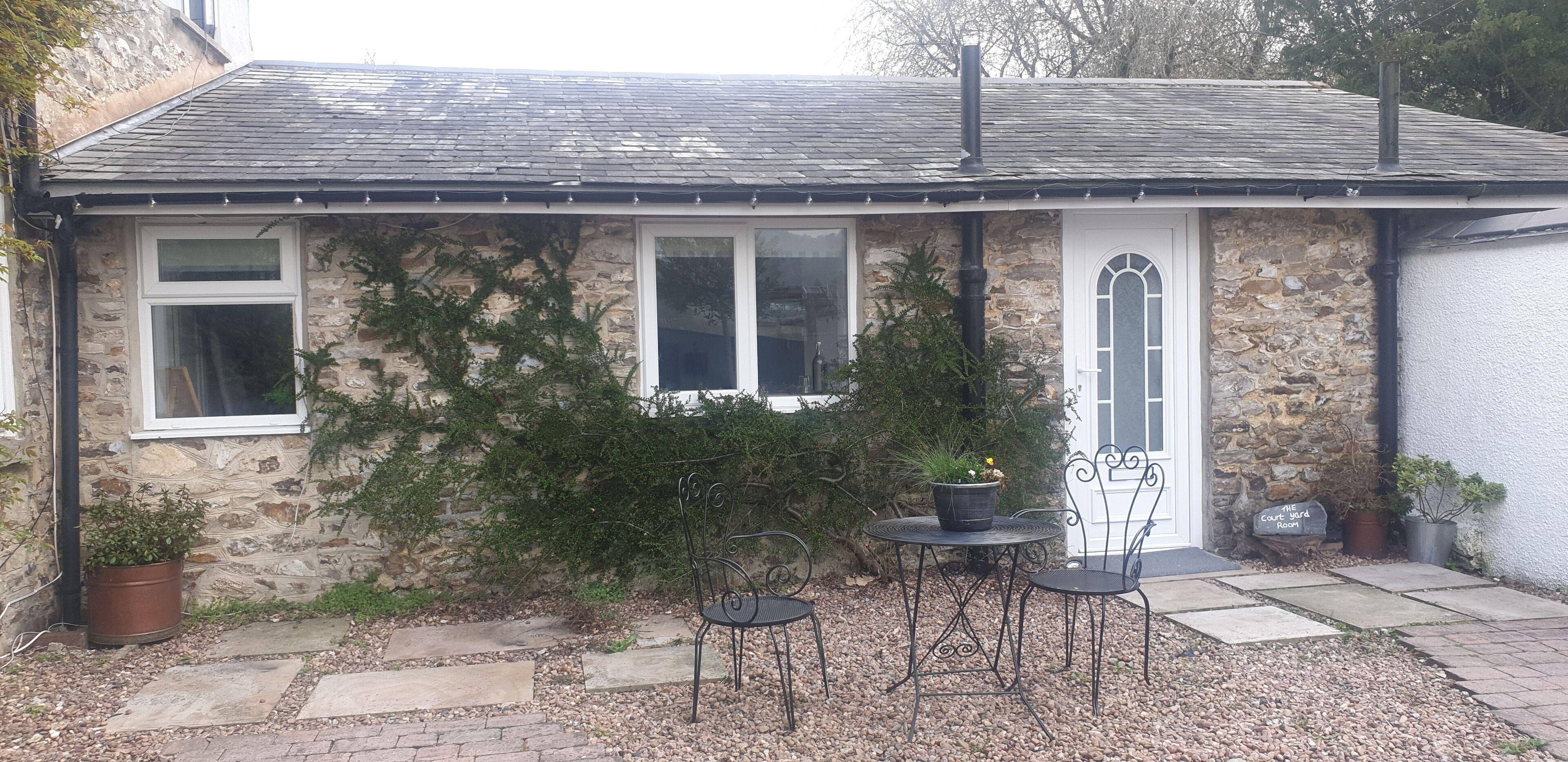 A stone house with a white door, two windows, and a small garden with a table and chairs.