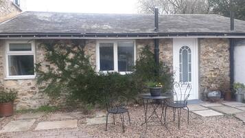A stone house with a white door, two windows, and a small garden with a table and chairs.