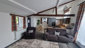 Un amplio espacio de sala con un sofá, un sillón y una mesa de comedor con sillas. Se ve una cocina con estufa y armarios al fondo.