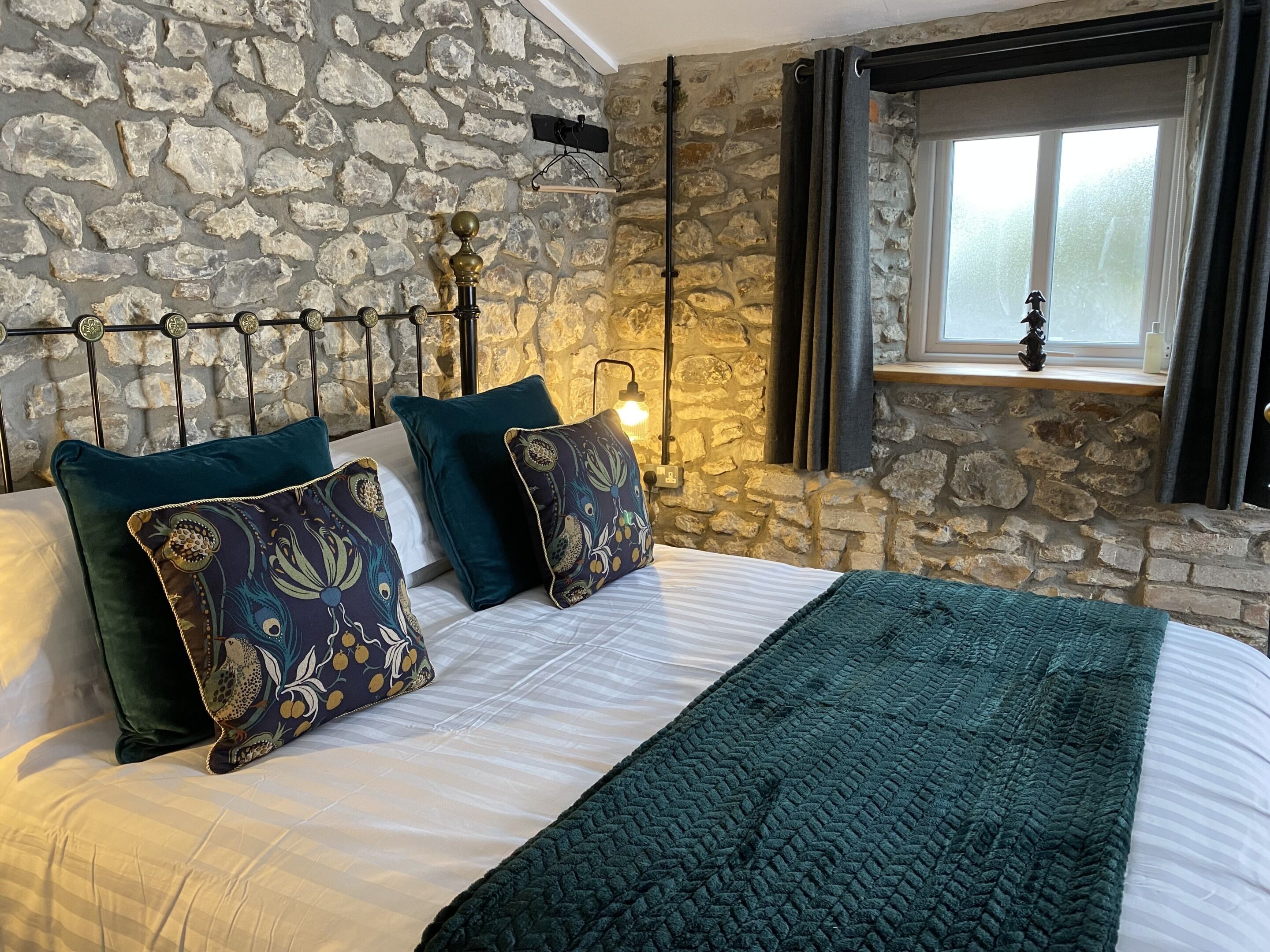 Un dormitorio con pared de piedra, cama con almohadas estampadas, colcha y ventana con cortinas.