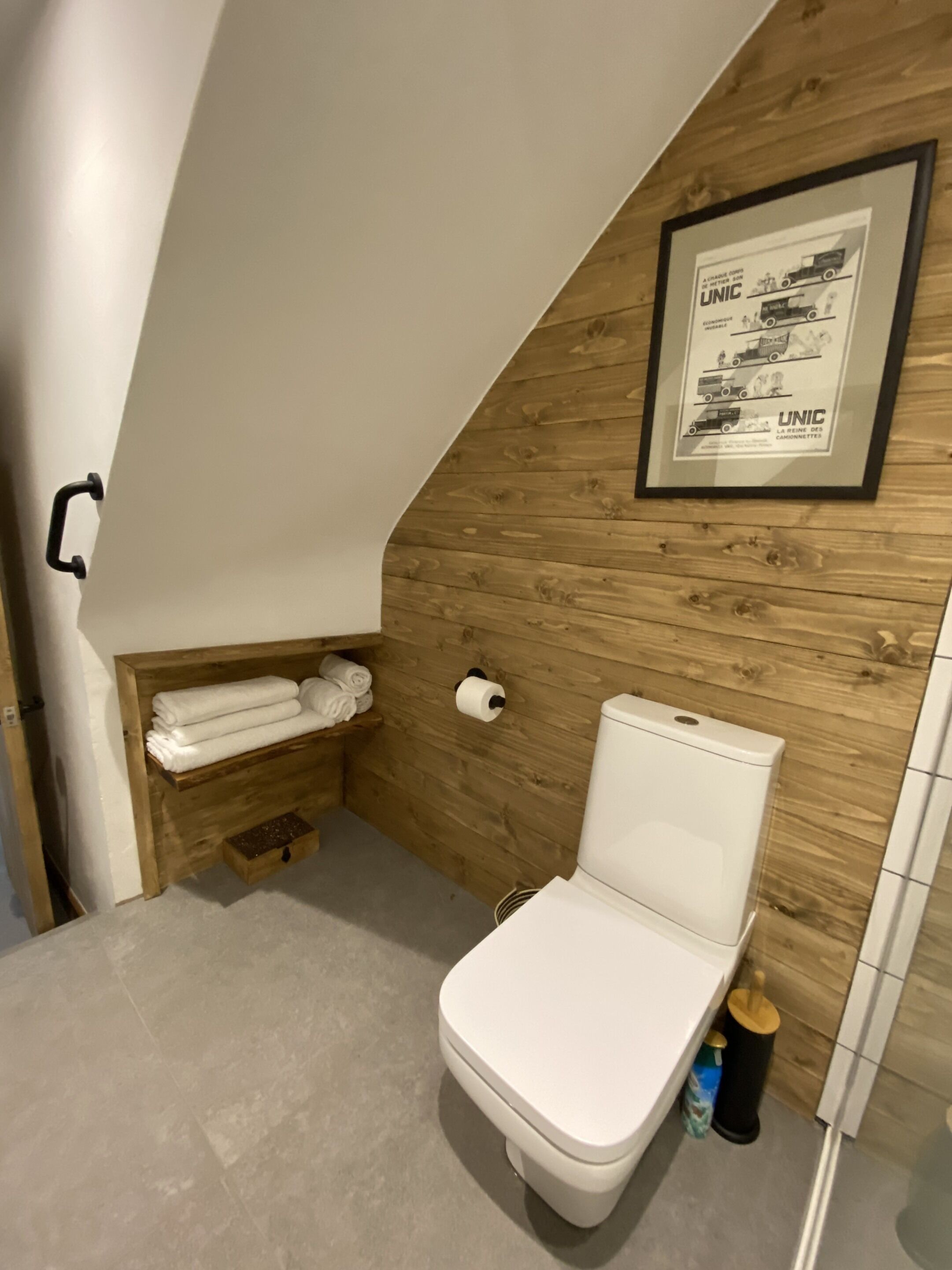 Une petite salle de bain aux murs inclinés, avec un mur en bois, des toilettes, une étagère avec des serviettes et une affiche encadrée.