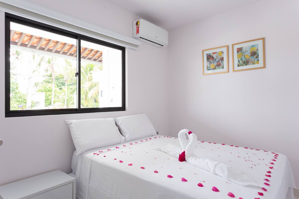 A hotel room with a bed, white bedding, a window with a view of greenery, and two framed pictures on the wall.