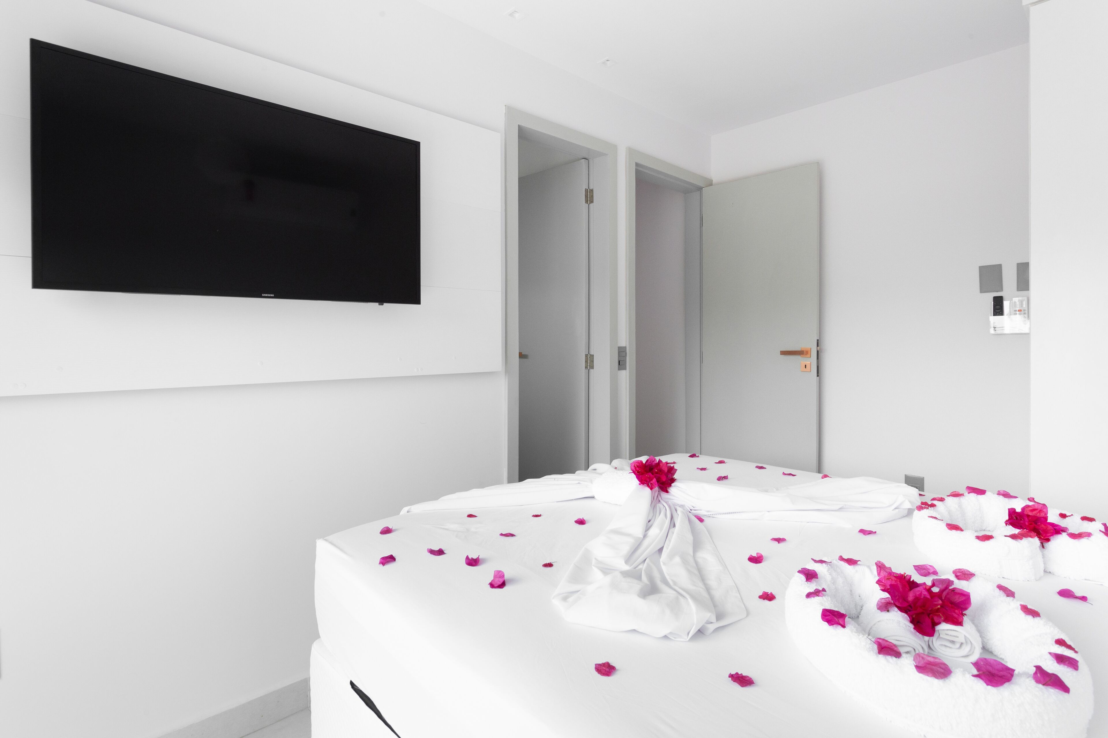 A modern hotel room with a large flat-screen TV mounted on the wall, a bed with white linens and rose petals, and a door leading to another room.