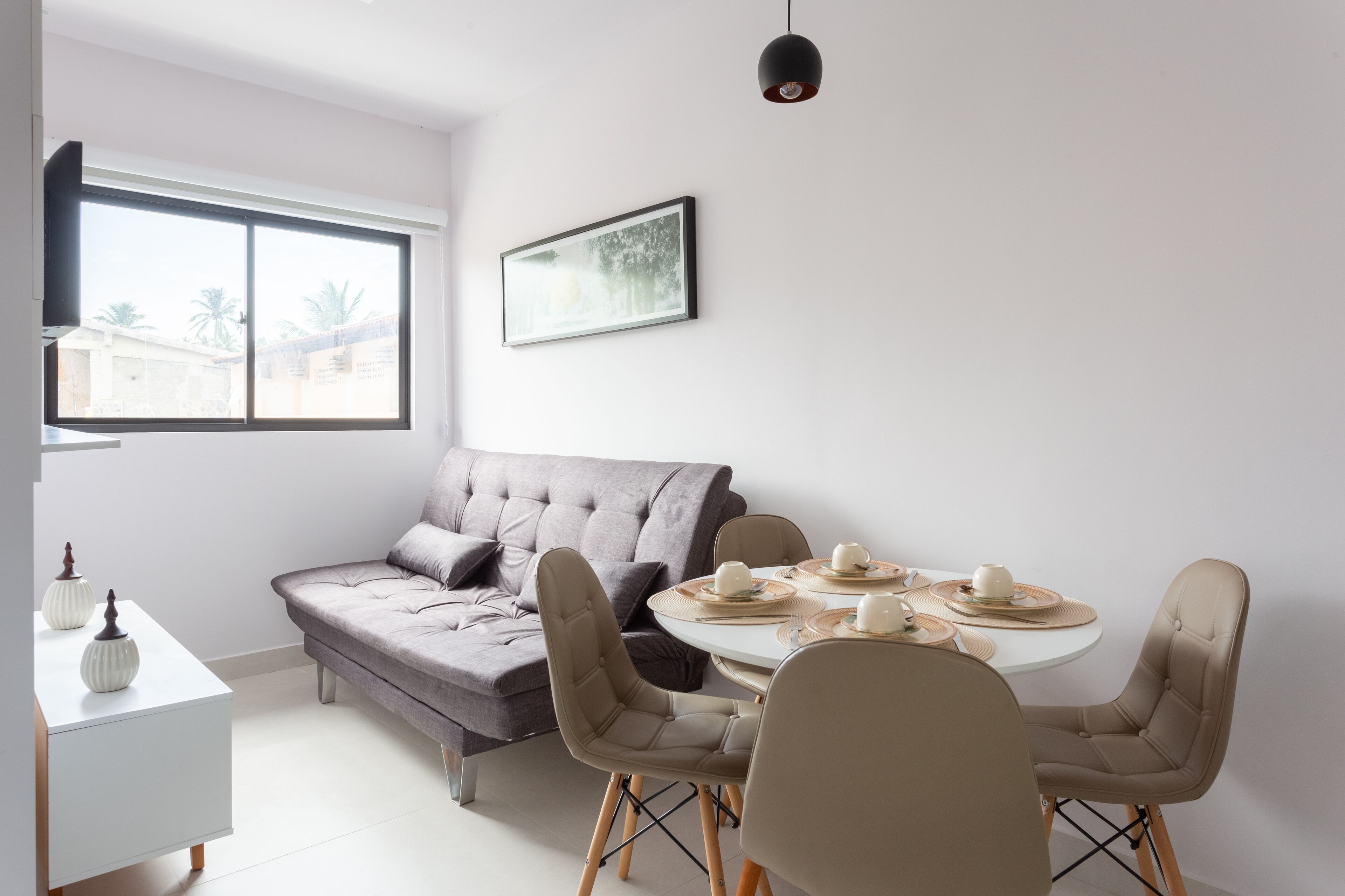 A modern living room with a grey sofa, a dining table with chairs, and a large window.