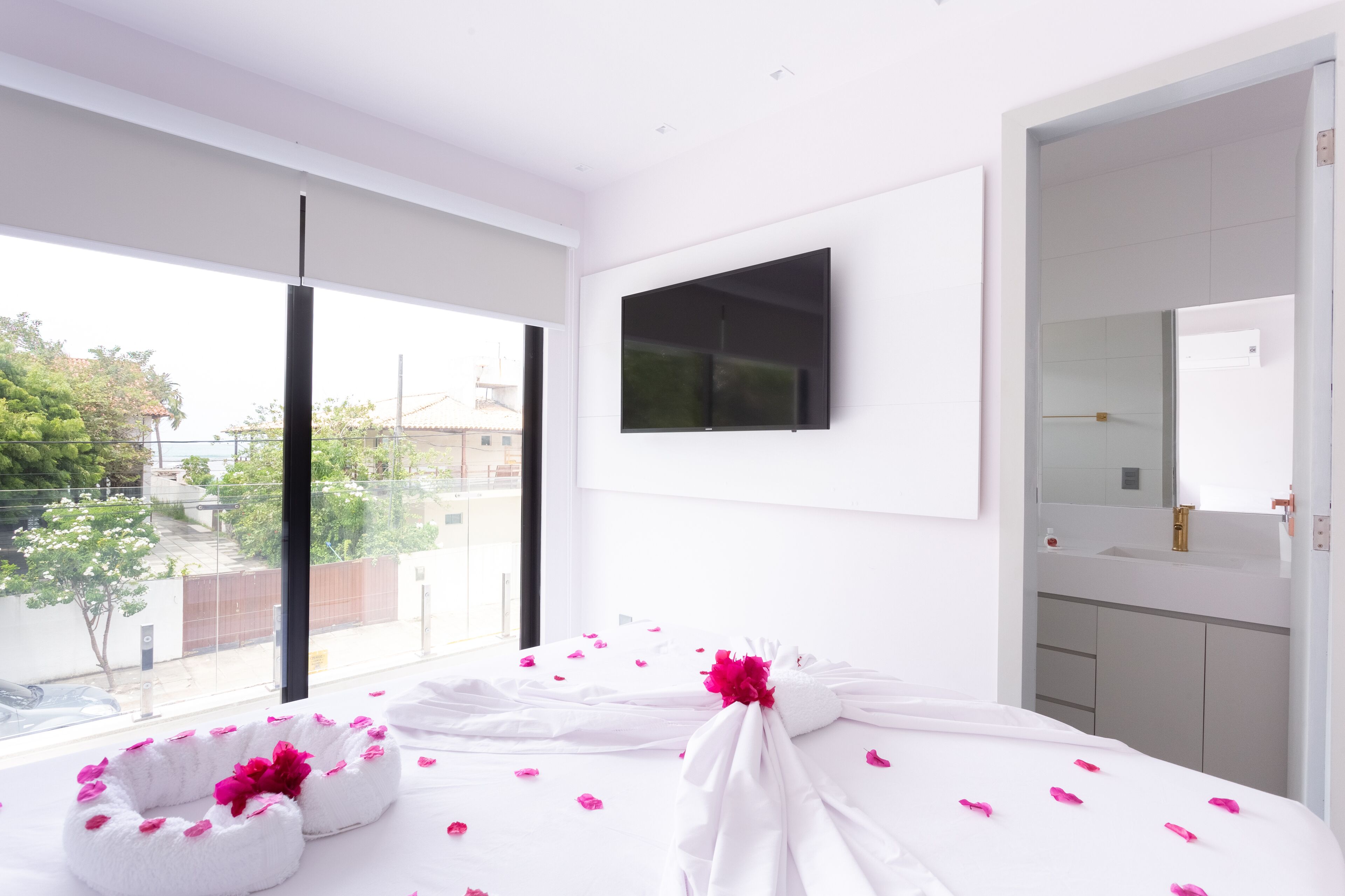 A modern hotel room with a large window, a flat-screen TV mounted on the wall, and a bathroom with a mirror and sink.