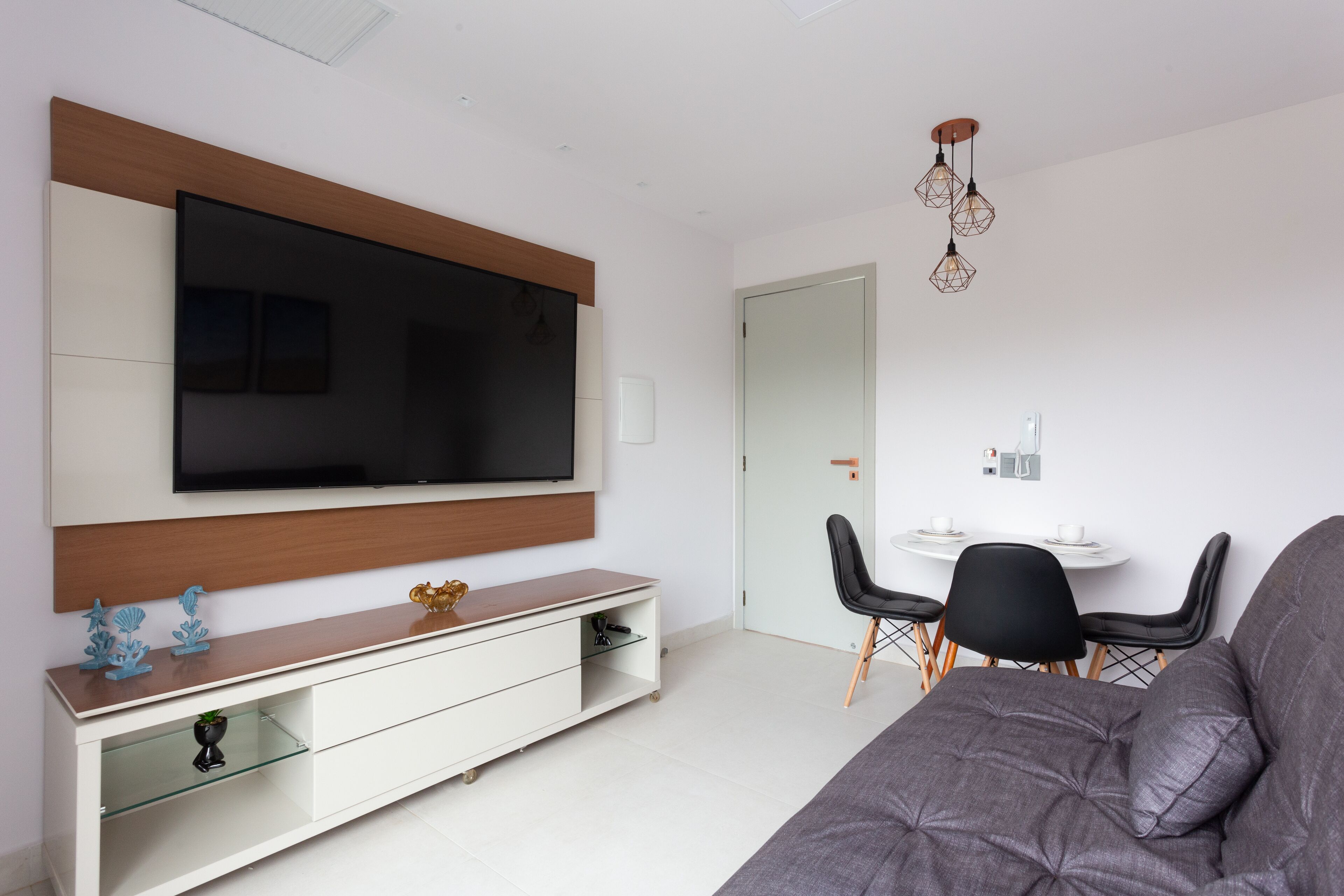 Una sala de estar moderna con un televisor de pantalla plana montado en un panel de madera, un mueble de televisión de vidrio con objetos decorativos, un sofá gris, una mesa de comedor con sillas y una lámpara colgante.