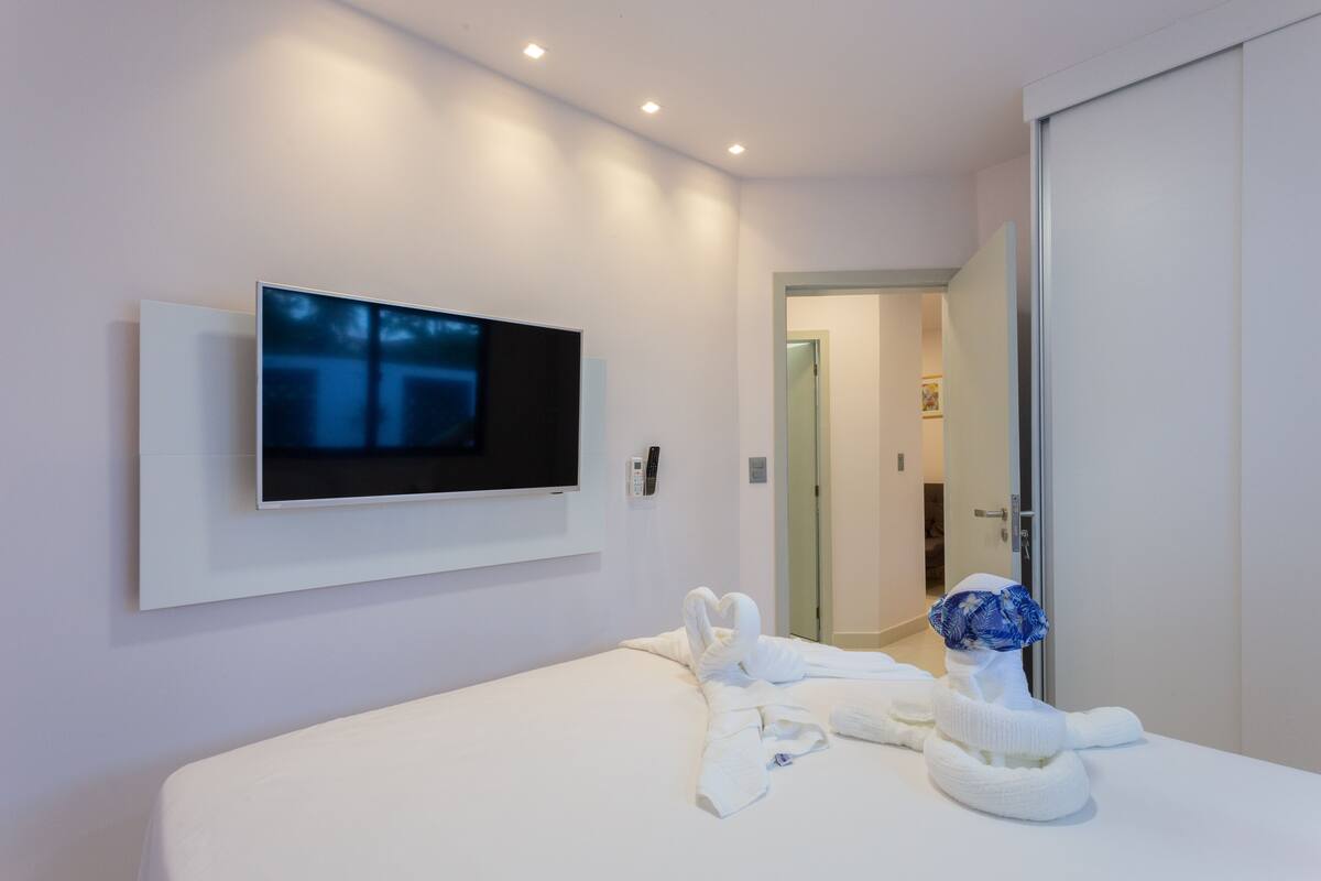 A modern hotel room with a flat-screen TV mounted on the wall, a neatly made bed with white linens, and a blue floral headscarf on the bed.