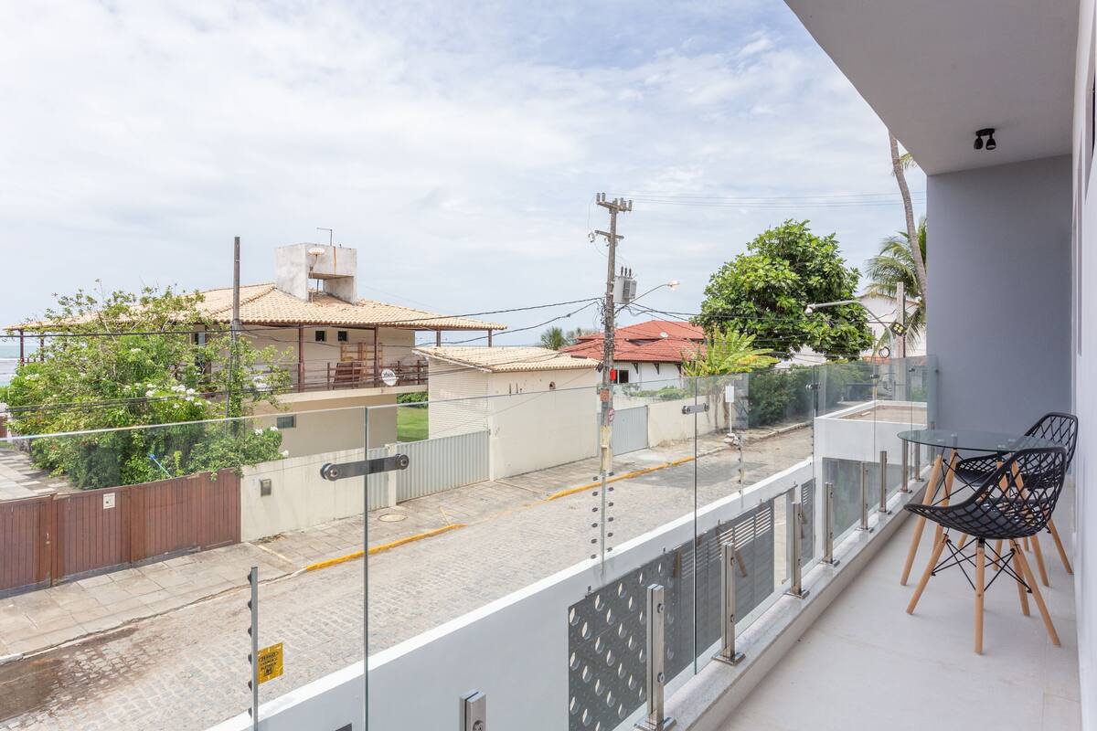 A balcony with a glass railing, a table with chairs, and a view of a street with buildings and trees.