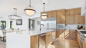 Une cuisine moderne avec des meubles en bois, un grand îlot central, des appareils électroménagers en acier inoxydable et des luminaires suspendus.